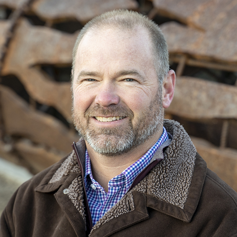 Kelley Mingus, a man in a brown jacket, is smiling in front of a rusty metal structure.